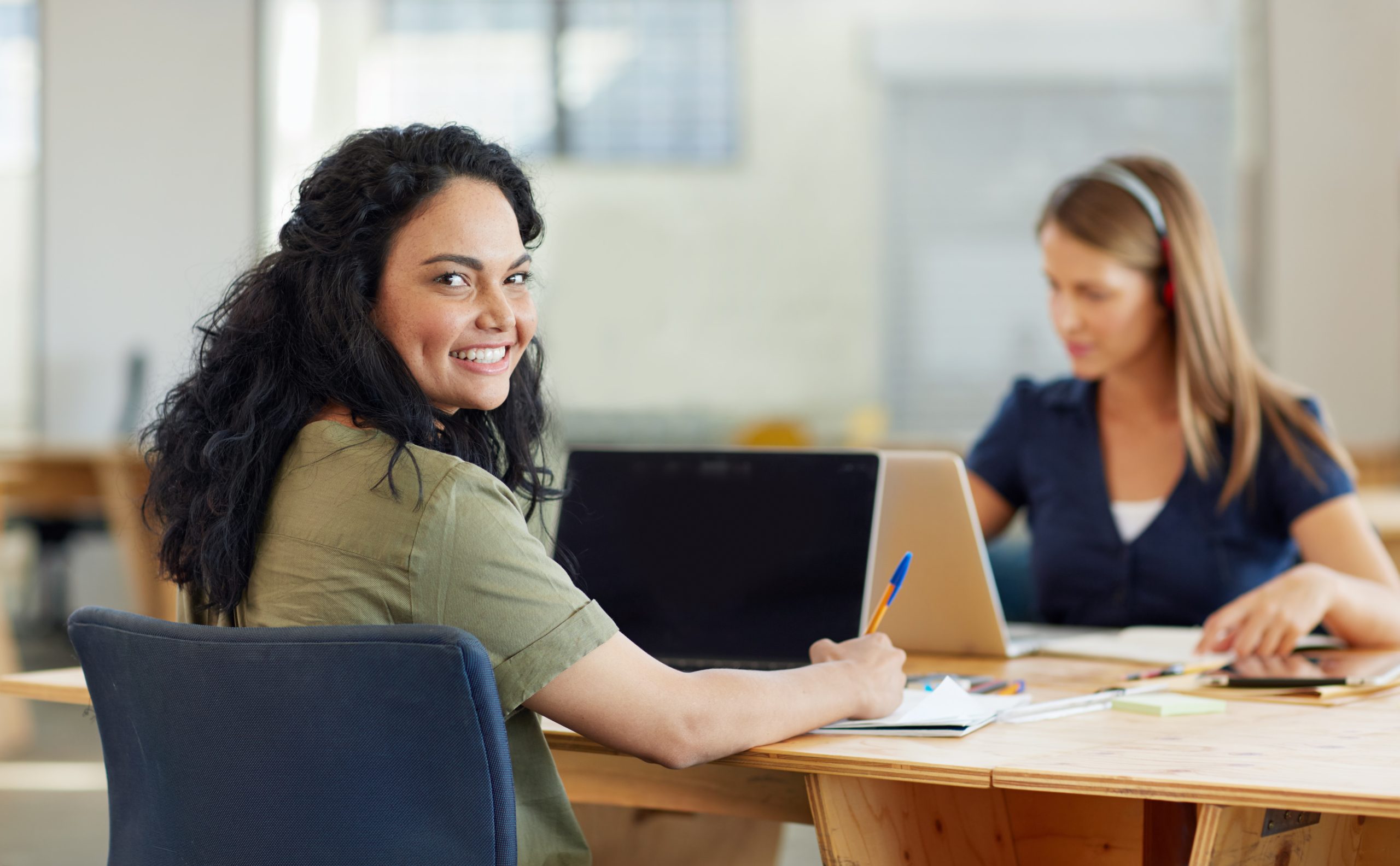 women at a desk working on a computer with another woman in the background on the computer listening to something with headphones