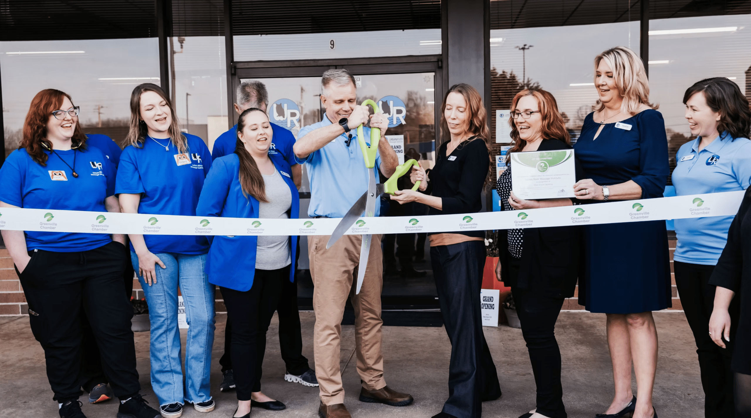 A group of staff and community members gather outside for a ribbon-cutting ceremony as a man cuts the ribbon with oversized scissors.